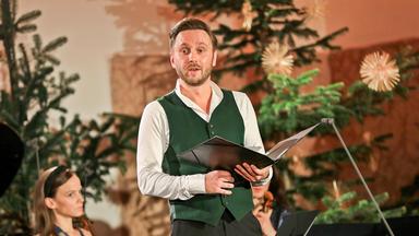 Das Bild zeigt einen Mann, der auf einer Bühne steht und anscheinend singt oder spricht. Er trägt ein grünes Weste über einem weißen Hemd und hat einen ernsten Gesichtsausdruck. In seiner linken Hand hält er ein schwarzes Musikheft. Im Hintergrund sind mehrere Tannenbäume dekoriert, die für eine festliche Atmosphäre sorgen. Auf der linken Seite des Bildes ist eine Frau mit einem Instrument, vermutlich einer Geige, zu sehen. Die gesamte Szene vermittelt eine besinnliche, weihnachtliche Stimmung, die in der Nikolauskirche in Bad Ischl aufgenommen wurde.