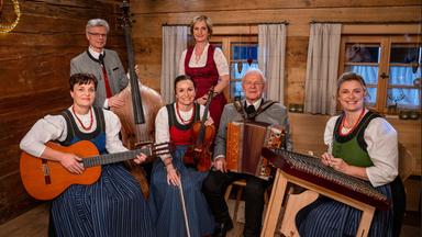 Das Bild zeigt eine Musikgruppe in einem rustikalen Raum mit Holzverkleidung. Im Vordergrund sitzen fünf Personen: vier Frauen und ein Mann. Die Frauen tragen traditionelle Trachtenkleider, die aus einer Kombination von farbigen Oberteilen und langen, gestreiften Röcken bestehen. Sie tragen auch Schmuck, der aus roten Perlen besteht.

Der Mann sitzt in der Mitte, gekleidet in ein graues Sakko mit traditionellem Schnürverschluss, und spielt ein Akkordeon. Eine Frau hält eine Gitarre, während eine andere eine Violine in der Hand hat. Neben dem Mann steht eine weitere Frau, die ein Hackbrett spielt. Im Hintergrund ist ein großes Fenster mit Vorhängen zu sehen, durch das sanftes Licht fällt, sowie einige Dekorationen. Der Boden ist aus Holz und der Raum wirkt warm und einladend. 

Diese Szene vermittelt eine festliche und traditionelle Atmosphäre, die mit der Musik und den Trachten verbunden ist.