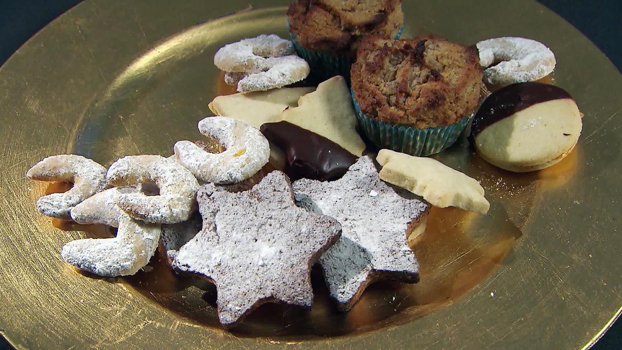 Das Bild zeigt eine Auswahl von traditionellen Weihnachtskeksen, die auf einem runden, goldenen Teller angerichtet sind. Die Kekse sind in verschiedenen Formen und Farben dargestellt. Unter ihnen befinden sich:  - Sauber ausgestochene, sternförmige Kekse in einem dunklen Farbton, bestäubt mit Puderzucker. - Helle, hornförmige Kekse und einige Ringkekse, die ebenfalls mit Puderzucker bestreut sind. - Ein paar Kekse mit Schokoladenglasur. - Außerdem sind kleine, runde Muffins in einer blauen Papierform sichtbar.  Der Kontrast zwischen den hellen und dunklen Keksen sowie die glatte Oberfläche des goldenen Tellers verleihen dem Arrangement eine festliche Ausstrahlung, passend zur Adventszeit in Österreich.