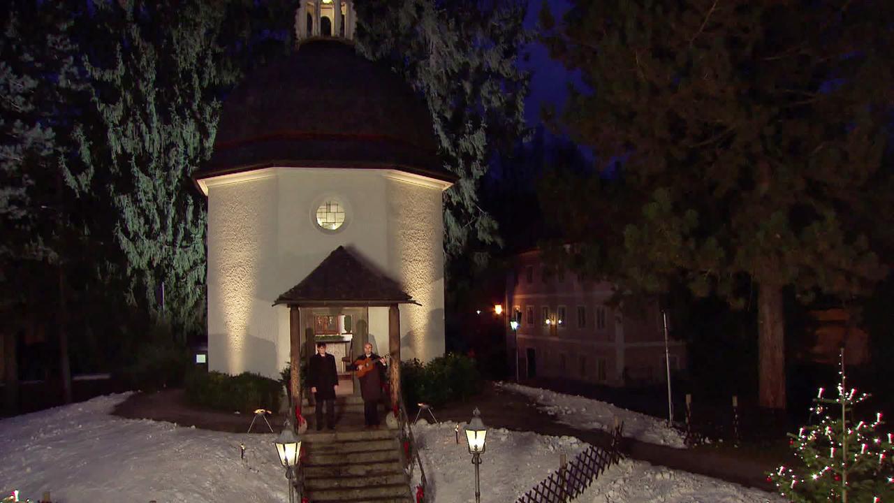 Das Bild zeigt die Gedächtniskapelle in Oberndorf, Österreich, in der Abenddämmerung. Die Kapelle hat eine runde Form mit einem aufwendig gestalteten, schrägen Dach, das zwischen den Bäumen hervorragt. Vor der Kapelle führt eine kurze Treppe zu einem Eingang, der mit zwei Holzsäulen flankiert ist. Das Gebäude ist in sanftem Licht erleuchtet, was einen warmen, einladenden Eindruck vermittelt.  Im Vordergrund stehen zwei Personen, die musizieren. Ein Musiker hält eine Gitarre, während der andere möglicherweise singt oder eine andere Instrumentation spielt. Um die Kapelle herum ist der Boden mit Schnee bedeckt, und es sind einige festliche Lichter aufgestellt. Auf der linken Seite sind mehrere Laternen sichtbar, die sanftes Licht abgeben. Im Hintergrund sind weitere Gebäude sichtbar, die in gedämpften Farben gehalten sind. Die Atmosphäre ist ruhig und festlich, was auf die Adventszeit hinweist.