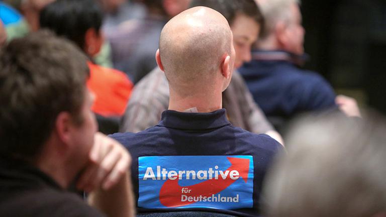 Glatzköpfiger Mann im Sitzen von hinten mit Schild auf dem Rücken "Alternative für Deutschland". 
