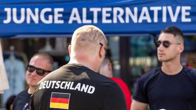 Rückansicht eines kahl rasierten Mannes mit der Aufschrift Deutschland auf seinem T-Shirt vor einem Stand der Jungen Alternativen.