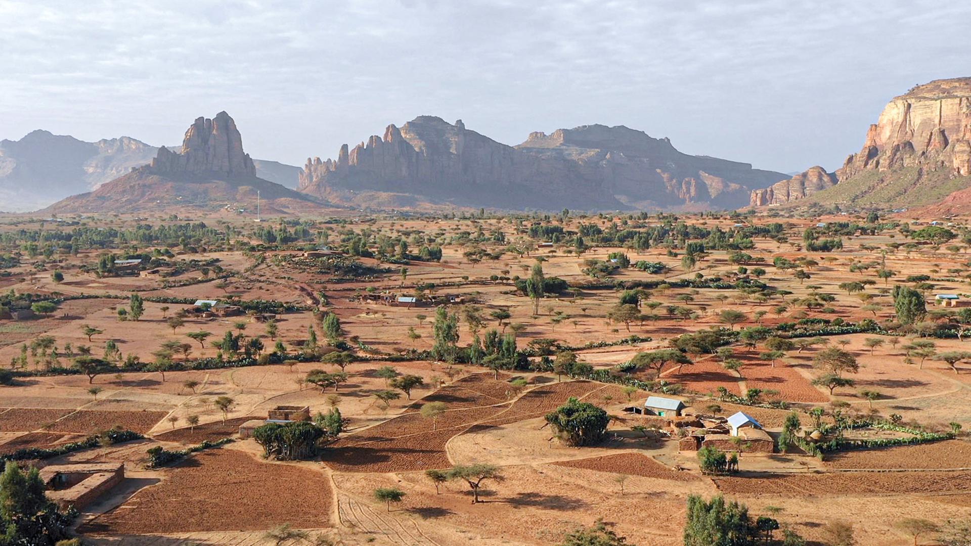 Karge Landschaft mit wenigen Bäumen und Häusern. Im Hintergrund Berge.