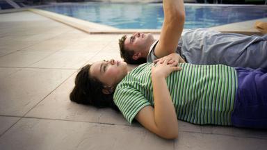 Sophie (Frankie Corio) und ihr Vater Callum (Paul Mescal) liegen auf dem Rücken auf einem gepflasterten Boden, im Hintergrund ist die blaue Wasseroberfläche eines Pools zu sehen.