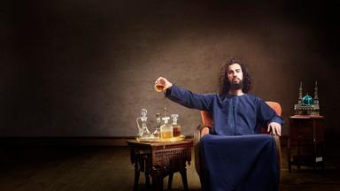 Omar sitzt in seiner traditionell muslimischen Kleidung auf einem Sessel und kippt die Flüssigkeit aus einem Whiskeyglas aus.