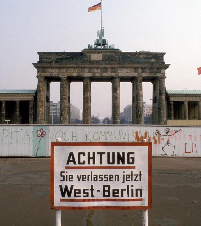 Blick auf das Brandenburger Tor hinter der Berliner Mauer. Im Vordergrund steht ein Schild mit der Aufschrift "Achtung! Sie verlassen jetzt West-Berlin".