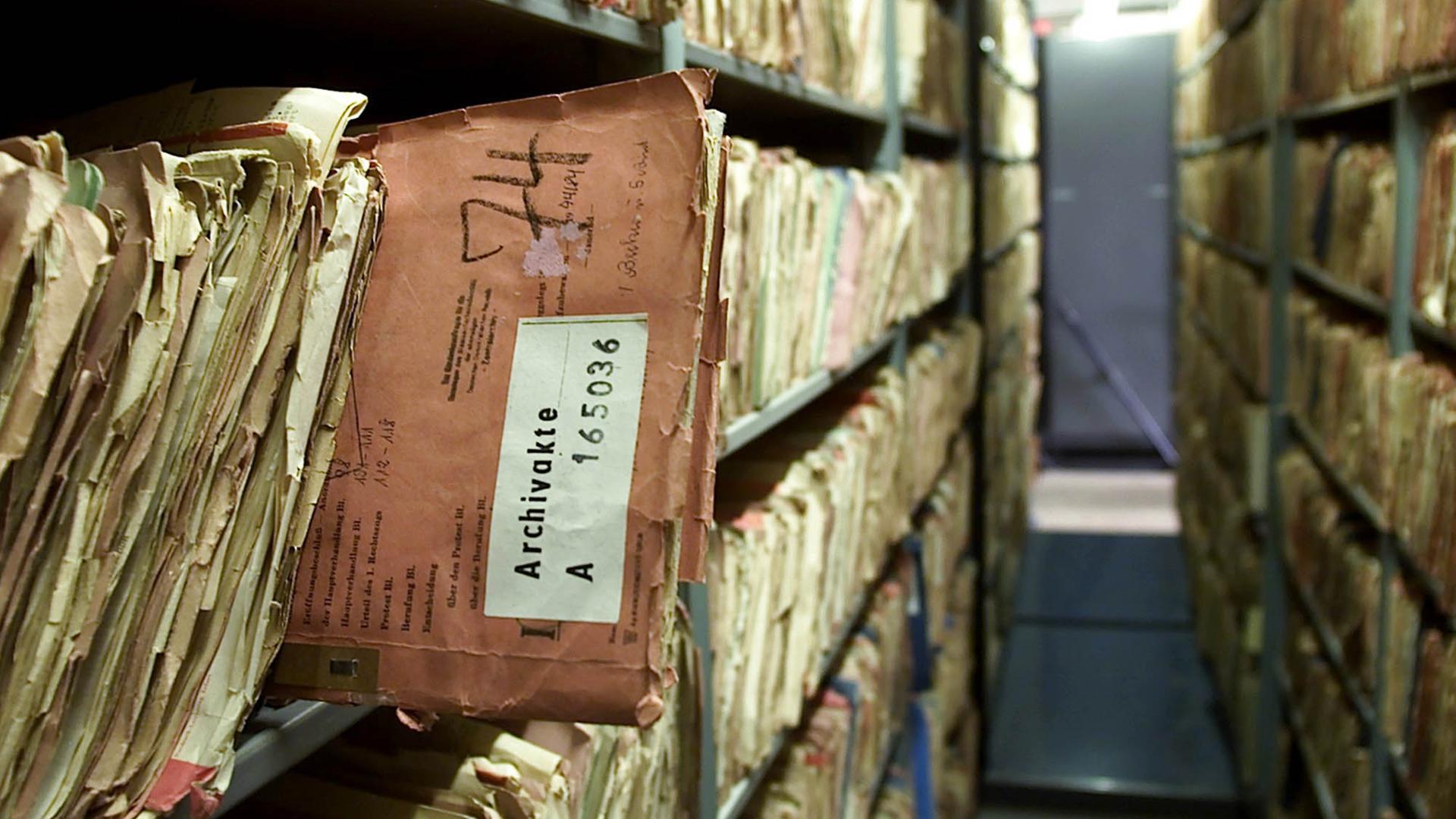 Dicht gefüllte Archivregale mit alten Akten. Im Vordergrund ragt eine abgegriffene, rötlich-braune Akte aus dem Regal.