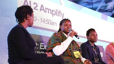 Auf einem Podium sitzten mehrere schwarze Personen. Eine Frau in buntem afrikanischem Gewand spricht in ein Mikrofon. Im Hintergrund wird der Schriftzug AI 2 Amplify eingeblendet.