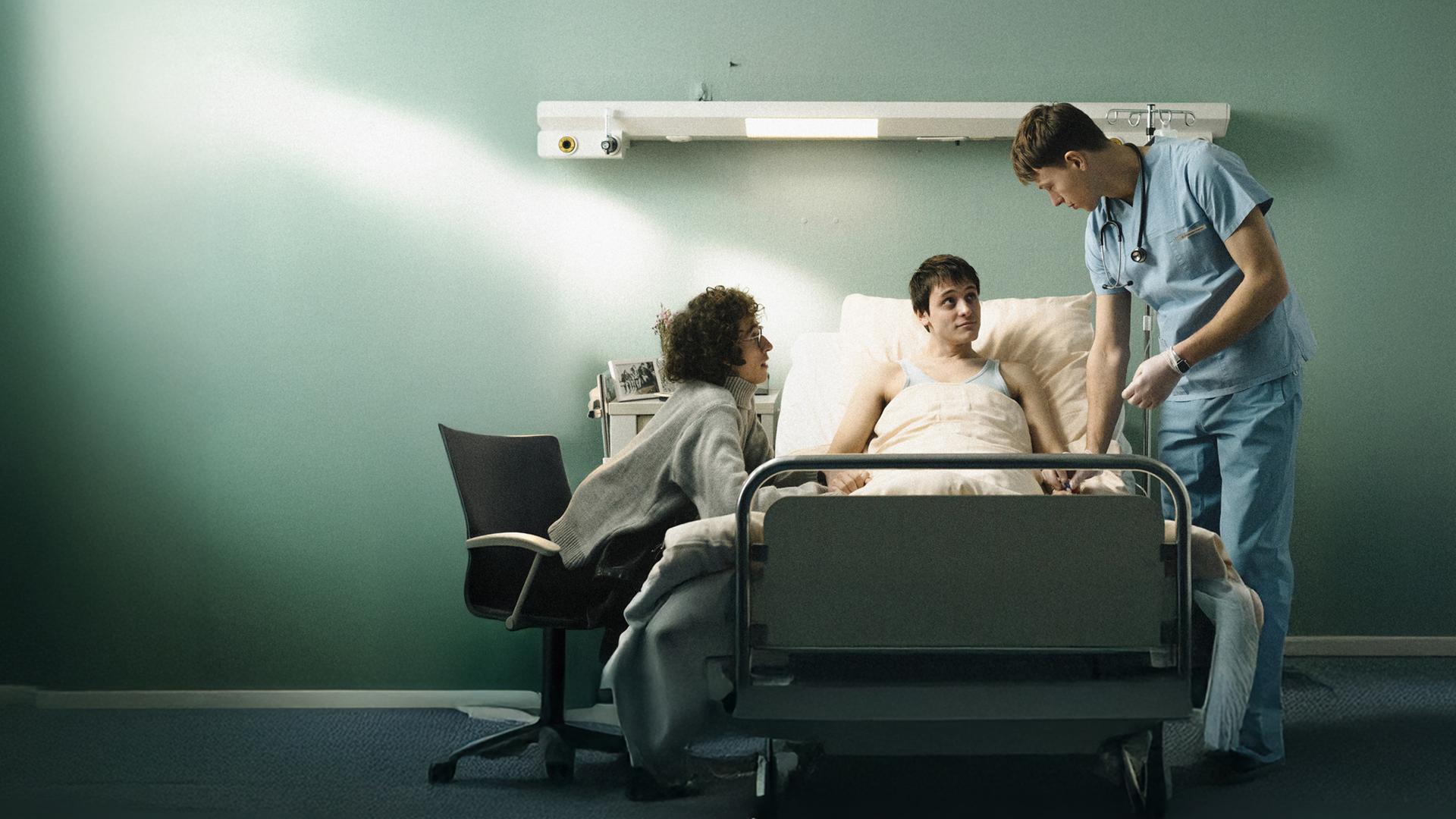 Dirk (Evangelos Tsarkowistas) am Bett - er hält Heikos Hand,. Auf der anderen Seite steht der Krankenpfleger Bernhard (Clemens Bobke).