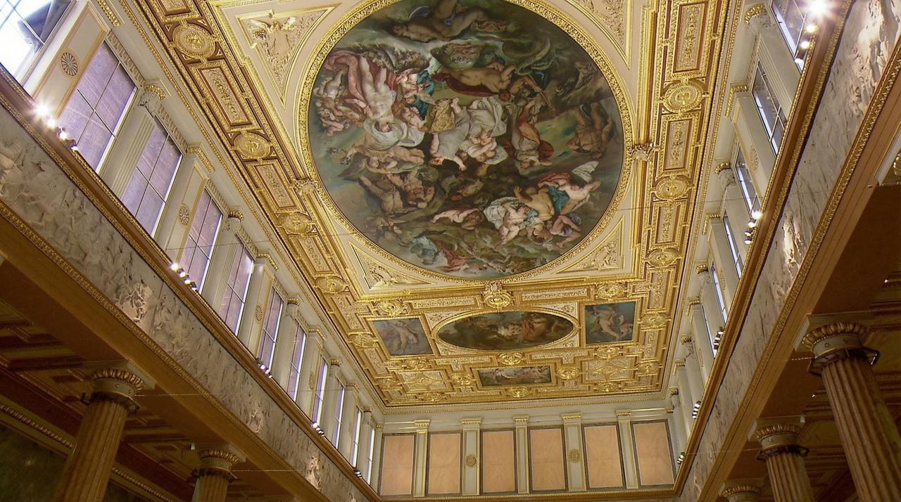 Das Bild zeigt die beeindruckende Inneneinrichtung der Akademie der bildenden Künste in Wien. Der Blick ist nach oben gerichtet und zeigt detaillierte Deckenmalereien, die in kräftigen Farben und aufwendigen Motiven gestaltet sind. Umrahmt von filigranem Goldverzierungen, sind mythologische Szenen und Figuren dargestellt.   Die Wände des Raumes sind mit eleganten, hellen Farben und strukturierten Elementen ausgestattet. Zierleisten und kunstvolle Reliefs sind sichtbar, die den historischen Charme des Gebäudes unterstreichen. Große Fenster an den Seiten sorgen für eine helle und freundliche Atmosphäre im Raum.  Die Architektur weist auf klassische Stilelemente hin, mit hohen Säulen, die eine majestätische Wirkung erzeugen. Insgesamt vermittelt das Bild einen Eindruck von Kunst und Geschichte, die in diesem bedeutenden kulturellen Umfeld zusammenkommen.