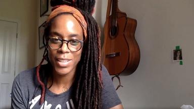 Eine Frau mit schwarzen langen Rastazöpfen sitzt vor einer Wand, an der Gitarren hängen.