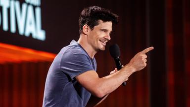 Alain Frei steht in der Mitte des Bildes. Er ist von der linken Seite zu sehen und streckt den Zeigefinger seiner rechten Hand nach vorne. 