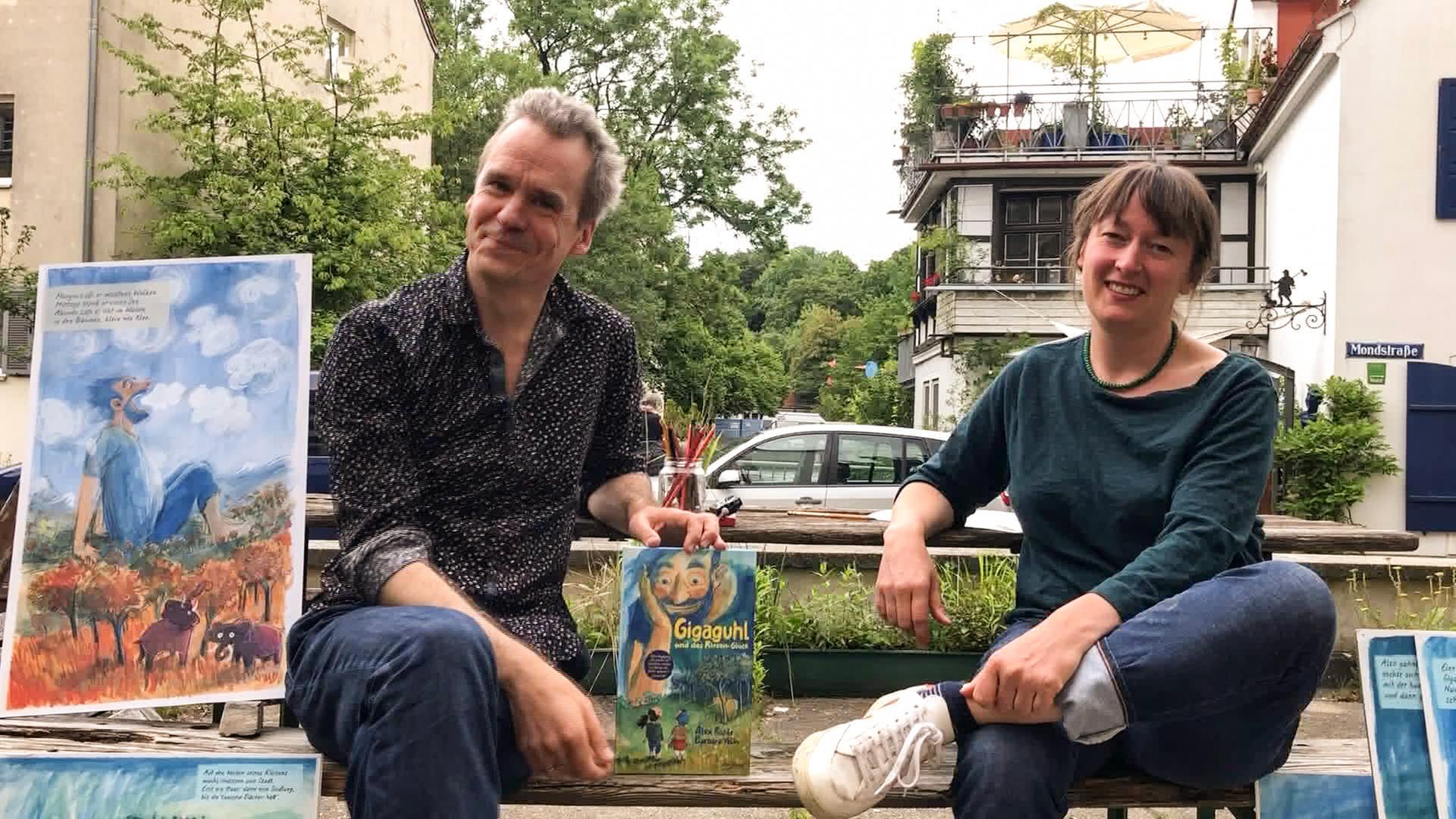 Ein Mann und eine Frau sitzen auf einer Bank im Freien und zeigen ein Kinderbuch und Zeichnungen aus dem Buch.