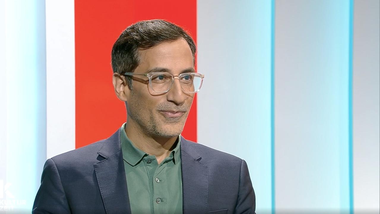 Ein lächelnder Mann mit Brille sitzt im Kulturzeit-Studio.