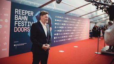 Ein Mann im Anzug steht auf einem roten Teppich zu einem Mikrofon gewandt. 