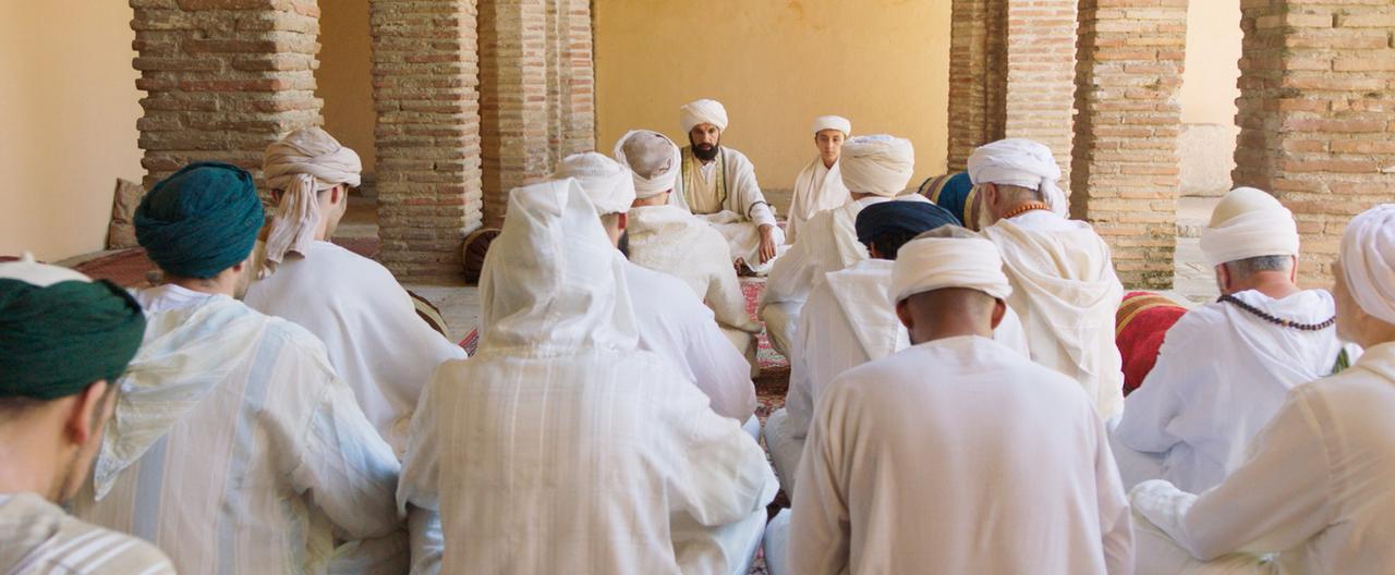 Das Bild zeigt eine Gruppe von Männern, die in einem Innenraum sitzen. Die meisten von ihnen tragen traditionelle, helle Roben und Kopfbedeckungen, während einige auch dunkle Kopfbedeckungen tragen. Der Raum wirkt historisch und ist durch Säulen mit unregelmäßig geformten Steinen strukturiert, die offensichtlich Teil der Architektur sind. Im Hintergrund sind einige dekorative Teppiche zu sehen, die auf dem Boden ausgelegt sind. Die Gesichter der sitzenden Männer sind größtenteils nicht sichtbar, da sie sich um einen zentralen Punkt versammeln. Ein Mann in der Mitte könnte eine herausragende Rolle einnehmen; er sitzt auf einer Art erhöhter Plattform und scheint zu sprechen oder zu leiten. Der Raum hat eine warme, beige Wandfarbe und strahlt eine ruhige Atmosphäre aus.