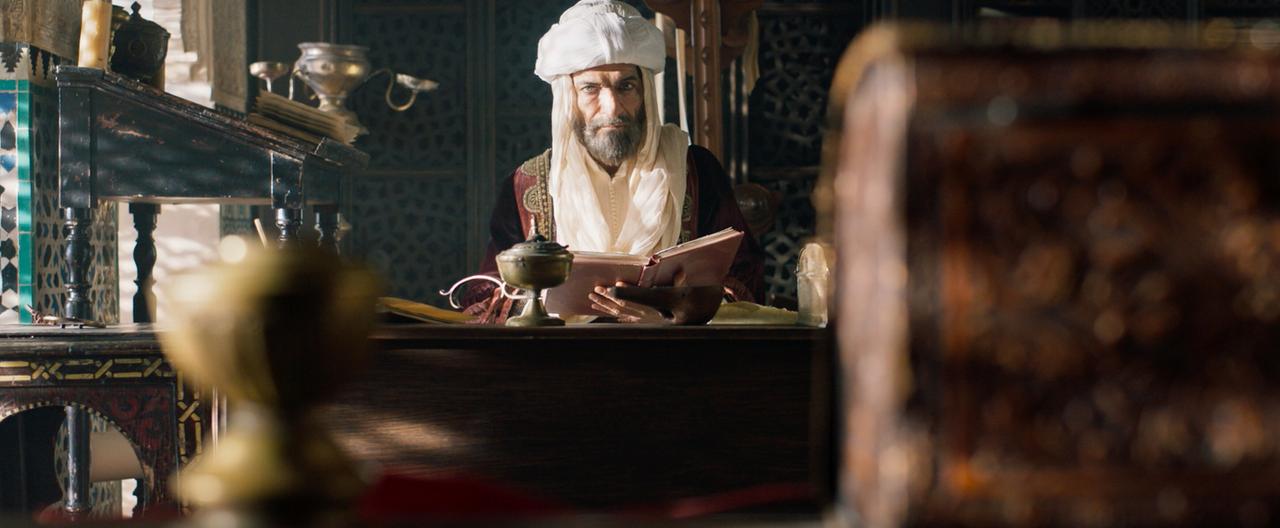 Das Bild zeigt einen Mann, der an einem Tisch sitzt. Er trägt traditionelle arabische Kleidung, bestehend aus einem langen, roten Gewand und einem weißen Kopftuch. Sein Gesicht ist von einem langen Bart umrahmt, und er hält ein Buch in der Hand. Der Hintergrund ist detailreich mit ornamentalem Design, das inspirierend und historisch wirkt. Auf dem Tisch sind verschiedene Gegenstände zu sehen, darunter eine dekorative Schale, ein Schreibutensil und ein paar andere unbekannte Objekte. Das Licht fällt sanft durch einen Fensterbereich, wodurch eine warme und einladende Atmosphäre entsteht. Die gesamte Szene vermittelt ein Gefühl von Weisheit und Historie.