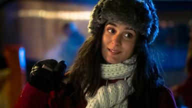 Eine junge Frau mit langen dunklen Haaren steht draußen bei Nacht. Sie trägt eine graue Fellmütze, einen dicken weißen Strickschal und einen roten Wintermantel. In der Hand hält sie einen Skistock, ihr Gesichtsausdruck wirkt nachdenklich und leicht lächelnd.