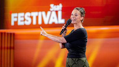 Kabarettistin steht auf der Mitte der Bühne. In ihrer rechten Hand hält sie ein Mikrofon. Mit der linken Hand zeigt sie mit einem Finger nach vorne. Der Hintergrund ist rot und orange beleuchtet.