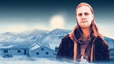 Ein Mann mit langen, blonden Haaren, Bart und Kapuzenpullover im Porträt. Im Hintergrund sind einstöckige Häuser vor einem Gebirge zu sehen.