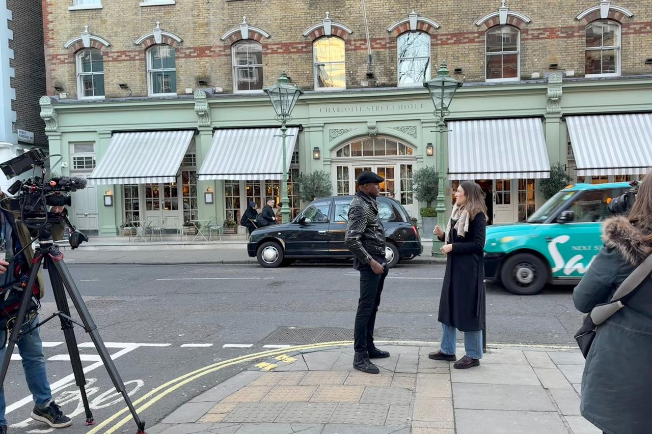 Das Bild zeigt eine Straßenszene in London. Im Vordergrund befinden sich zwei Personen, die ein Gespräch führen. Die erste Person ist ein Mann, der eine schwarze Lederjacke mit Nieten, schwarze Hose und schwarze Schuhe trägt und eine schwarze Mütze aufhat. Die zweite Person ist eine Frau in einem langen, schwarzen Mantel mit einem grauen Schal und blauen Jeans.   Im Hintergrund ist das "Charlotte Street Hotel" zu sehen, ein Gebäude mit hellgrünem Mauerwerk und weißen Streifenmarkisen über den Fenstern. Es gibt eine Reihe von Tischen und Stühlen vor dem Hotel, die darauf hinweisen, dass es ein Restaurant gibt.   Am Straßenrand steht ein schwarzes Londoner Taxi. Im Bild ist auch ein Kameramann zu sehen, der seine Kamera auf einem Stativ platziert hat, um das Interview aufzuzeichnen. Es scheint, als ob der Mann beim Interview eine Art Love Coach ist, während die Frau wahrscheinlich die Korrespondentin ist.