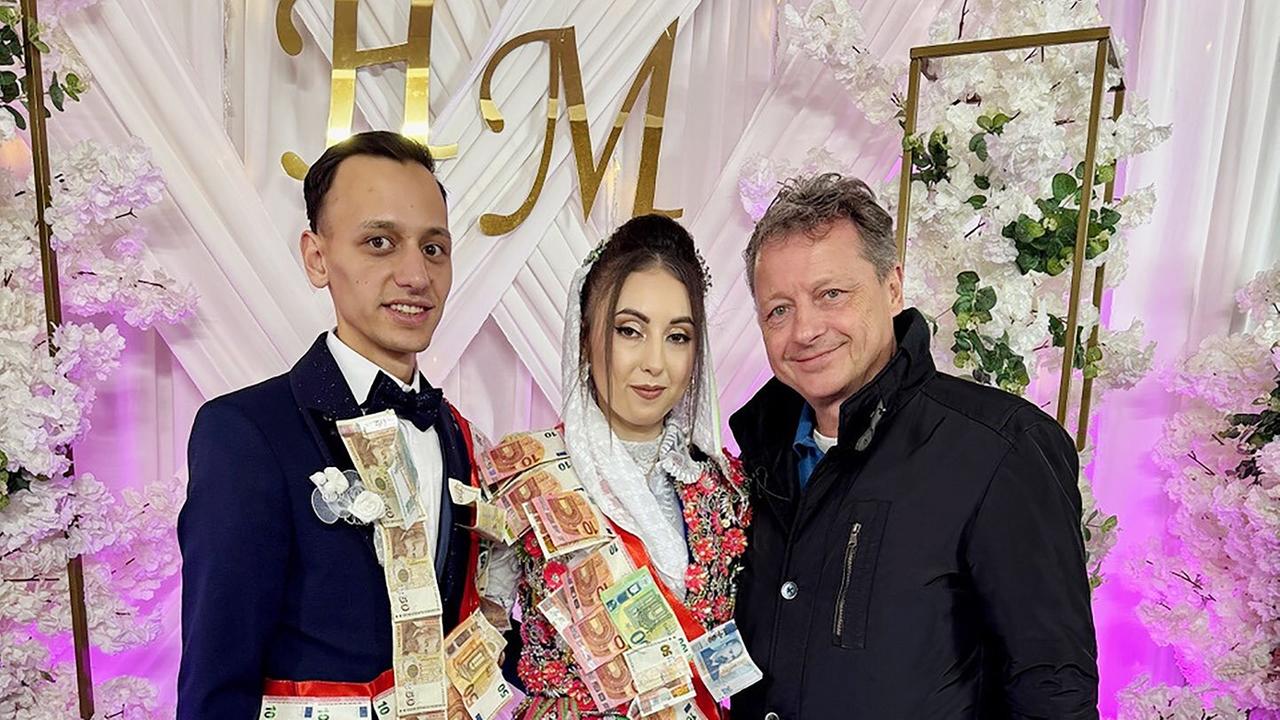 Das Bild zeigt eine Hochzeitsfeier in einem festlichen Moment. Zwei Personen, ein Bräutigam und eine Braut, stehen in der Mitte und lächeln. Der Bräutigam trägt einen eleganten Anzug mit einer Fliege und hat Geldscheine über die Brust drapiert. Die Braut trägt ein traditionelles, bunt verziertes Kleid mit Blumenmustern und hat ebenfalls Geldscheine an ihrer Kleidung angebracht. Sie trägt eine weiße Kopfbedeckung, die auf Hochzeitsbräuche hinweist.  Im Hintergrund ist eine dekorierte Kulisse mit großen, weißen Stoffbahnen und einem goldenen Schriftzug „HM“ sichtbar. Zudem sind künstliche Blumenarrangements zu sehen, die den Raum festlich schmücken. Die Umgebung hat eine farbige Beleuchtung, die dem Ambiente eine warme und feierliche Atmosphäre verleiht. Rechts von den beiden Hochzeitsgästen steht ein älterer Herr, der ebenfalls lächelt und in einem schwarzen Mantel gekleidet ist. Der Boden ist gefliest.   Das Bild vermittelt die fröhliche Stimmung einer traditionellen Hochzeit und zeigt die kulturellen Bräuche, die mit solchen Feierlichkeiten verbunden sind.