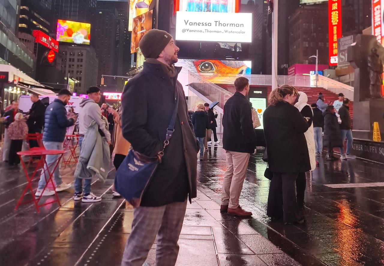 Das Bild zeigt eine belebte Straßenszene in New York City, insbesondere im Bereich von Times Square. Es ist Nacht, und der Boden ist nass, wahrscheinlich von einem kürzlichen Regen, was zu einem glitzernden Reflexionseffekt auf dem Straßenbelag führt.   Im Vordergrund steht ein Mann mit Bart und einer Mütze, der in dunkler Kleidung gekleidet ist. Er wirkt nachdenklich und schaut nach oben auf die bunten, leuchtenden Werbetafeln und Bildschirme, die mit verschiedenen Farben und Bildern beleuchtet sind.   Um ihn herum stehen mehrere andere Personen, die ebenfalls auf ihre Handys schauen oder in die Richtung der Lichter blicken. Diese Personen sind divers in Bezug auf ihre Kleidung, von Winterjacken bis hin zu lässiger Freizeitkleidung. In der Mitte des Bildes sind zwei rote Klapptische und Stühle zu sehen, die typisch für die Struktur von Times Square sind, wo Menschen oft verweilen.   Im Hintergrund sind hohe Gebäude sichtbar, die die Skyline von New York prägen, und es gibt Werbetafeln, die Informationen oder Werbung präsentieren. Der gesamte Bereich ist lebhaft und vermittelt eine Atmosphäre von urbanem Leben und Energie.