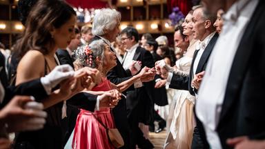 Das Bild zeigt eine Tanzszene während des Opernballs 2026. In einem festlich dekorierten Saal, der elegant mit roten Vorhängen und Kristallleuchtern ausgestattet ist, befinden sich zahlreiche Ballgäste, die in Paaren tanzen.   Im Vordergrund sind mehrere Personen sichtbar, die sich an den Händen halten. Eine ältere Frau mit grauen Haaren und einer pinken, fließenden Kleid sieht fröhlich aus. Sie tanzt neben einer Frau in einem weißen, eleganten Kleid. Um sie herum sind auch andere Gäste in formeller Abendkleidung, darunter Herren in Smoking und Damen in formellen Ballkleidern.  Der Hintergrund zeigt eine Mischung aus weiteren Tänzern und einem erhöhten Bereich, wahrscheinlich eine Bühne, auf der möglicherweise eine musikalische Darbietung stattfindet. Der gesamte Raum vermittelt eine festliche und glamouröse Atmosphäre des Balls.
