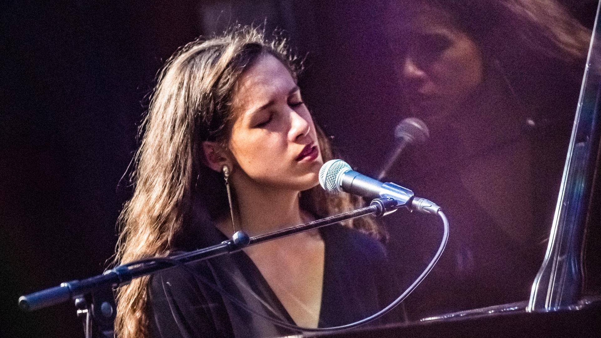 Musizierende Person am Mikrofon auf einer Bühne, beleuchtet von Bühnenlicht, mit sichtbarem Instrument und Performance-Situation.