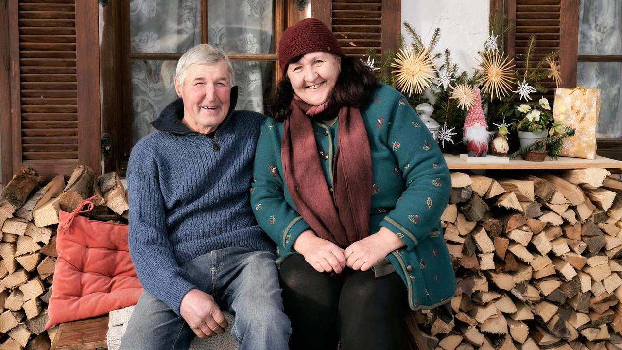 Auf dem Bild sitzen zwei Personen, ein Mann und eine Frau, eng beieinander auf einer Bank. Der Mann hat kurzes, graues Haar und trägt einen blauen Pullover, während die Frau dunkelbraunes Haar hat, das zu lockeren Wellen fällt. Sie trägt einen grünen Pullover mit bunten Mustern und einen roten Schal. Beide lächeln freundlich.  Im Hintergrund sind Holzfenster zu sehen, hinter denen Vorhänge mit einem floralen Muster hängen. Ein Holzstapel ist vor ihnen aufgeschichtet. Auf einer horizontalen Ablage hinter ihnen befinden sich einige weihnachtliche Dekorationen, darunter Sterne aus Stroh, ein kleiner Gnom und Geschenke.   Die Szene vermittelt eine gemütliche, winterliche Atmosphäre, die zu der geografischen Umgebung des Alpbachtals passt, in dem die winterlichen Landschaften und die Kultur thematisiert werden.