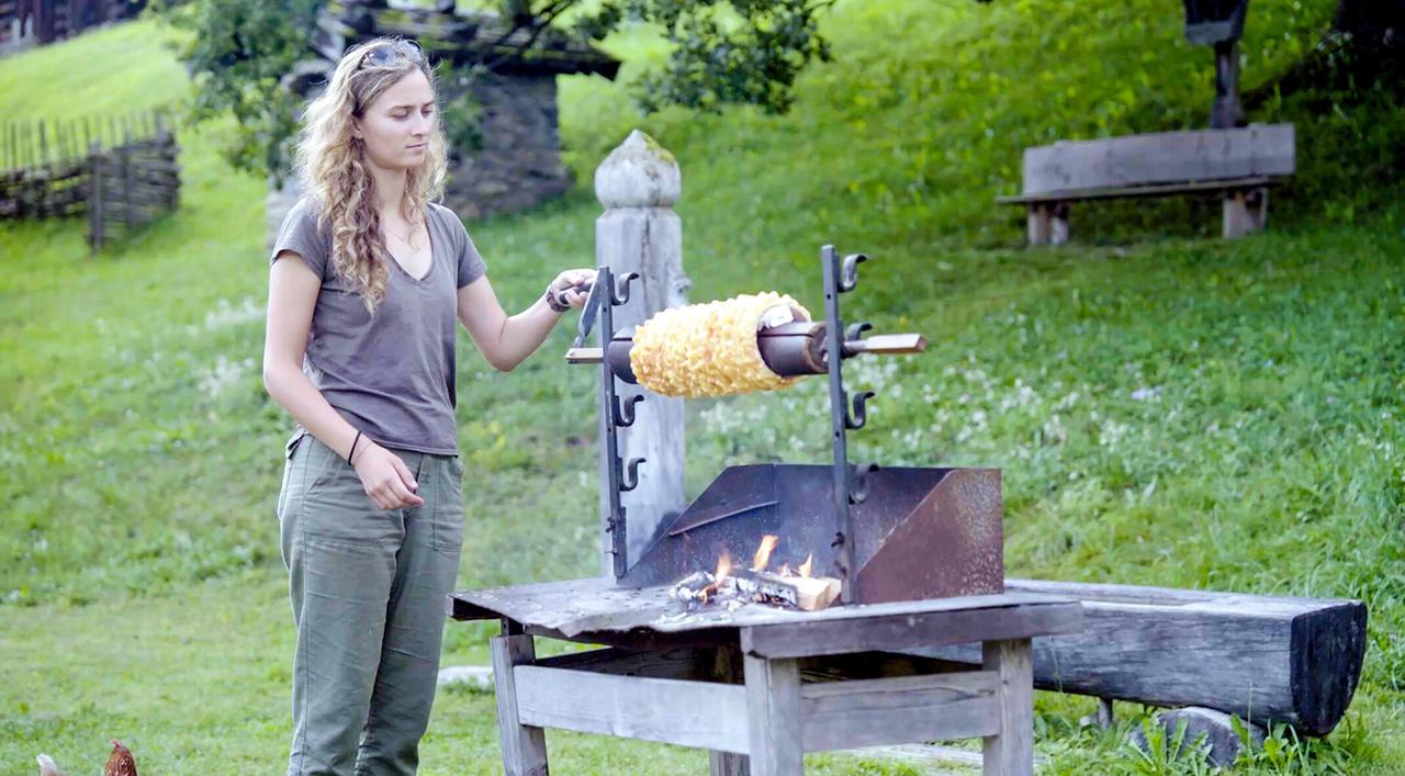 Das Bild zeigt eine junge Frau mit lockigem, blondem Haar, die an einem Holzstand steht, auf dem eine Prügeltorte zubereitet wird. Die Frau trägt ein graues T-Shirt und olivfarbene Hosen. Sie hält einen Stab in der Hand, mit dem sie die Prügeltorte, die auf einem langen Holzstab montiert ist, dreht. Unter dem Stand sieht man ein kleines Feuer, das brennt.   Im Hintergrund erstrecken sich grüne Wiesen mit einigen Bäumen und einem Zaun aus Holz. Auf dem Boden sind auch einige Hühner zu sehen. Das Bild vermittelt eine ländliche, alpine Atmosphäre, die Tradition und handwerkliches Geschick widerspiegelt.