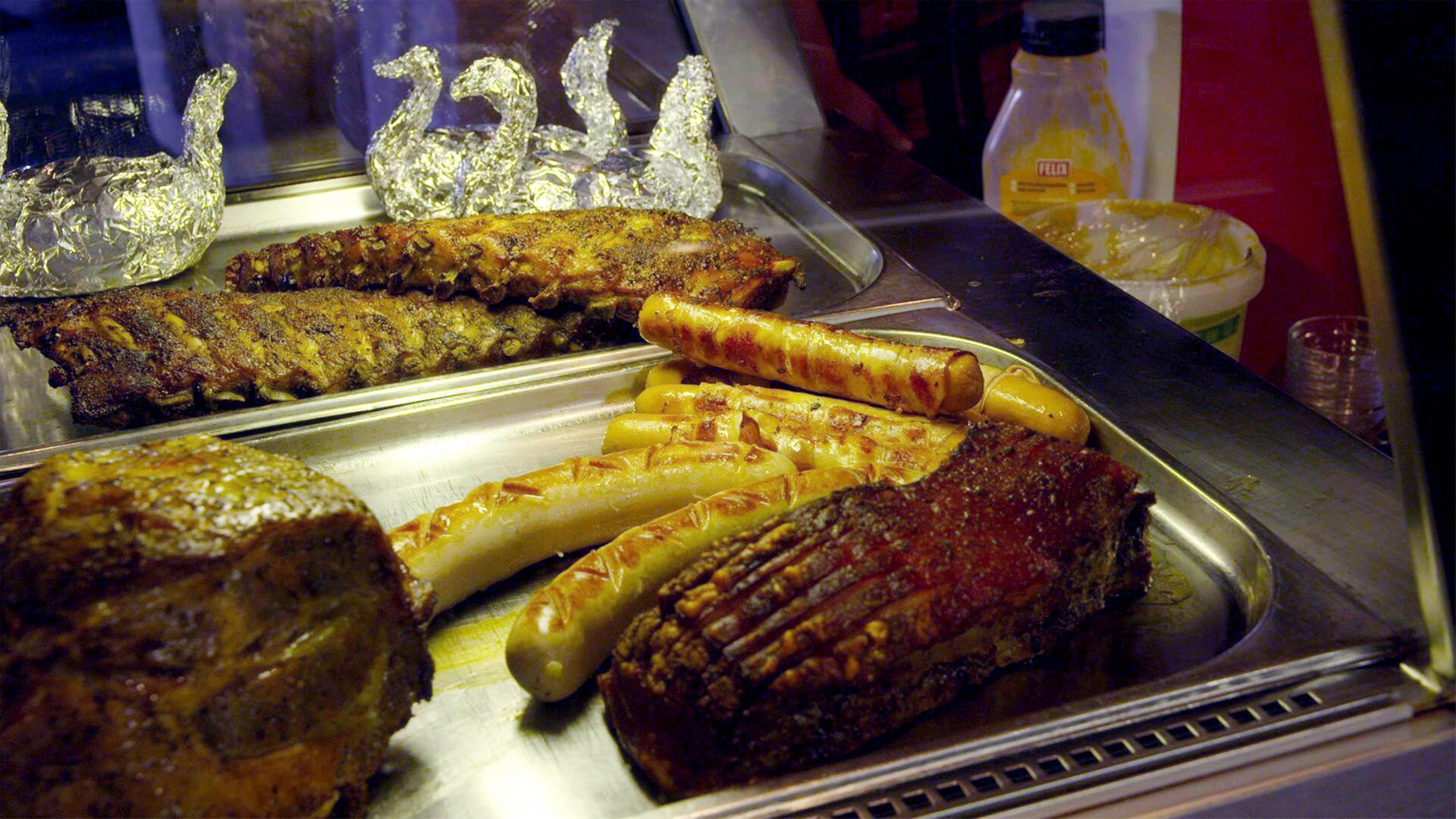 Das Bild zeigt eine Theke mit verschiedenen Grillgerichten. Im Vordergrund sind mehrere Fleischstücke und Würstchen auf einem rechteckigen Edelstahlblech angeordnet.   - Von links nach rechts sind sichtbar:   - Ein großes Stück Fleisch, wahrscheinlich Schweinebraten, mit einer glänzenden, leicht knusprigen Oberfläche und Rillen.   - Daneben liegt ein Rippchen, ebenfalls mit einer knusprigen Kruste.   - Vor dem Rippchen befinden sich mehrere Würstchen in verschiedenen Größen und Formen, einige scheinen gegrillt zu sein.    Hinter den Fleischgerichten sind zwei dekorative Schwäne aus Alufolie sichtbar, die die Präsentation auflockern. Im Hintergrund sind einige Flaschen mit gelben und weißen Flüssigkeiten zu erkennen, möglicherweise Saucen oder Dressing.  Die gesamte Szenerie wirkt einladend und typisch für eine gastronomische Einrichtung in den Bergen, wie sie in der Alpengastronomie zu finden ist.