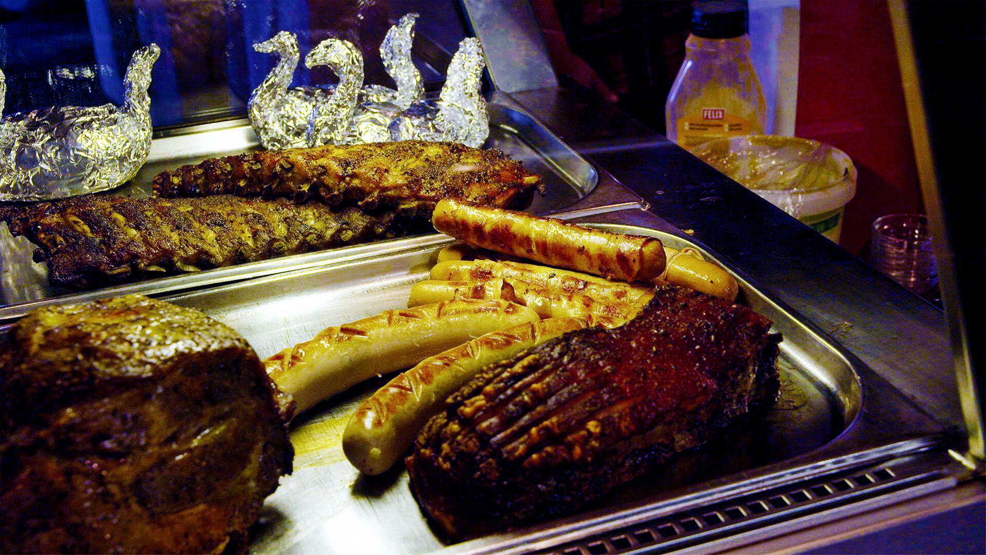 Das Bild zeigt eine Theke mit verschiedenen Grillgerichten. Im Vordergrund sind mehrere Fleischstücke und Würstchen auf einem rechteckigen Edelstahlblech angeordnet.   - Von links nach rechts sind sichtbar:   - Ein großes Stück Fleisch, wahrscheinlich Schweinebraten, mit einer glänzenden, leicht knusprigen Oberfläche und Rillen.   - Daneben liegt ein Rippchen, ebenfalls mit einer knusprigen Kruste.   - Vor dem Rippchen befinden sich mehrere Würstchen in verschiedenen Größen und Formen, einige scheinen gegrillt zu sein.    Hinter den Fleischgerichten sind zwei dekorative Schwäne aus Alufolie sichtbar, die die Präsentation auflockern. Im Hintergrund sind einige Flaschen mit gelben und weißen Flüssigkeiten zu erkennen, möglicherweise Saucen oder Dressing.  Die gesamte Szenerie wirkt einladend und typisch für eine gastronomische Einrichtung in den Bergen, wie sie in der Alpengastronomie zu finden ist.