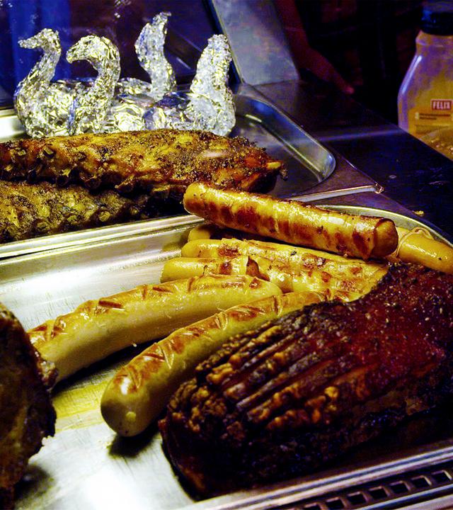 Das Bild zeigt eine Theke mit verschiedenen Grillgerichten. Im Vordergrund sind mehrere Fleischstücke und Würstchen auf einem rechteckigen Edelstahlblech angeordnet.   - Von links nach rechts sind sichtbar:   - Ein großes Stück Fleisch, wahrscheinlich Schweinebraten, mit einer glänzenden, leicht knusprigen Oberfläche und Rillen.   - Daneben liegt ein Rippchen, ebenfalls mit einer knusprigen Kruste.   - Vor dem Rippchen befinden sich mehrere Würstchen in verschiedenen Größen und Formen, einige scheinen gegrillt zu sein.    Hinter den Fleischgerichten sind zwei dekorative Schwäne aus Alufolie sichtbar, die die Präsentation auflockern. Im Hintergrund sind einige Flaschen mit gelben und weißen Flüssigkeiten zu erkennen, möglicherweise Saucen oder Dressing.  Die gesamte Szenerie wirkt einladend und typisch für eine gastronomische Einrichtung in den Bergen, wie sie in der Alpengastronomie zu finden ist.