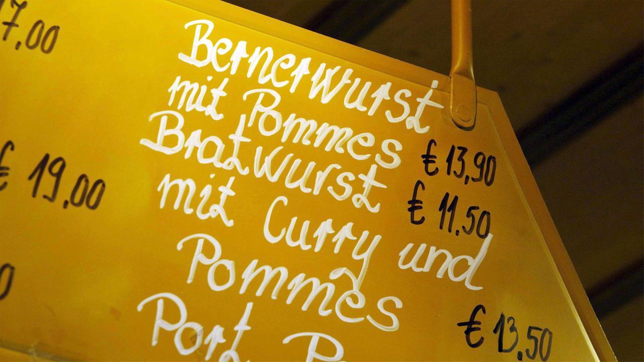 Das Bild zeigt eine Speisekarte, die an einem gelben Hintergrund hängt. Die Schrift ist in einer handgeschriebenen Art und Weise verfasst. Auf der Speisekarte stehen folgende Gerichte mit den entsprechenden Preisen in Euro:  1. Bernerwurst mit Pommes - 13,90 € 2. Bratwurst mit Curry und Pommes - 11,50 € 3. Port (eine weitere Speise) - 13,50 € 4. Ein weiteres Gericht oder eine Dienstleistung mit einem Preis von 19,00 €, der nicht genau benannt wird.  Der Text ist in verschiedenen Größen und Stilen gehalten, wobei die Preise farblich differenziert sind. Der gesamte Schriftzug vermittelt einen rustikalen und einladenden Eindruck, passend zur Alpengastronomie.
