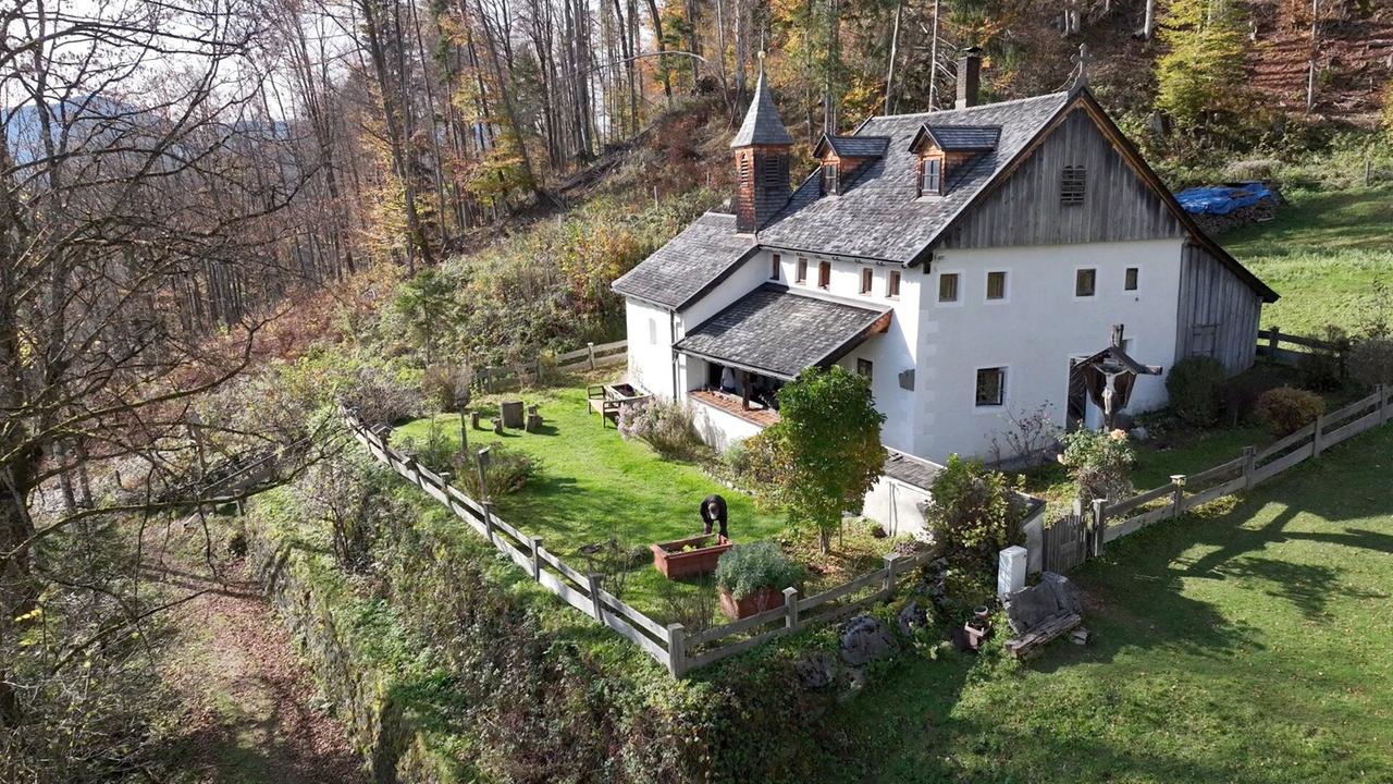 Das Bild zeigt die Einsiedlerkapelle in St. Johann, umgeben von einer malerischen Landschaft. Die Kapelle ist ein Gebäude im traditionellen alpinen Stil, mit einem schrägen Dach und einem kleinen Kirchturm, der an der Vorderseite aufragt. Sie ist weiß gestrichen mit Holzelementen, und es gibt mehrere Fenster, die Tageslicht hereinscheinen lassen.

Vor der Kapelle erstreckt sich ein gepflegter Garten mit grünem Rasen und bunten Blumen. Einige Sitzgelegenheiten befinden sich im Freien, darunter eine Bank aus Holz. In der Nähe sieht man einen Menschen, der in einem Gartenbeet arbeitet. Die Umgebung ist von Bäumen umgeben, die herbstlich gefärbt sind, und es sind einige weitere Waldgebiete im Hintergrund sichtbar. Das ganze Bild vermittelt einen ruhigen und harmonischen Eindruck, der die natürliche Schönheit der Region unterstreicht. 

Es gibt keine spezifischen Informationen zu bekannten Schauspielern oder Orten, da das Bild hauptsächlich die Kapelle und ihre Umgebung zeigt.