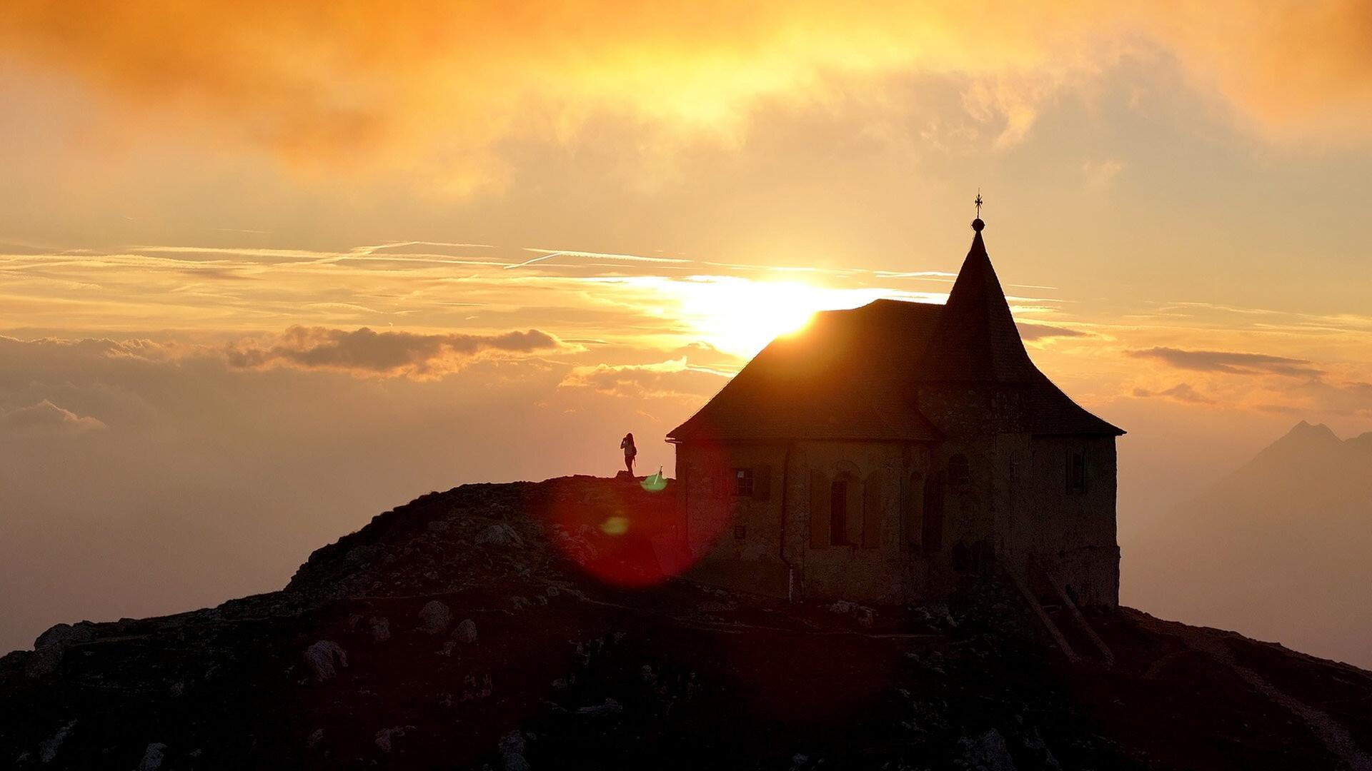 Das Bild zeigt die Dobratsch Deutsche Kapelle, eine Kirche auf einem Berggipfel in den Alpen. Die Kapelle hat einen spitzen Turm und ist im Hintergrund vor einem dramatischen Sonnenuntergang positioniert, bei dem orangefarbene und goldene Töne den Himmel füllen. Der Sonnenstrahl scheint hinter der Kapelle hervor und erzeugt eine leuchtende Silhouette. Im Vordergrund ist ein felsiger Hang zu sehen, und auf diesem Hang steht eine Person, die möglicherweise die Aussicht genießt. Die umgebenden Bereiche sind von Wolken umgeben, was dem Bild eine mystische Atmosphäre verleiht.