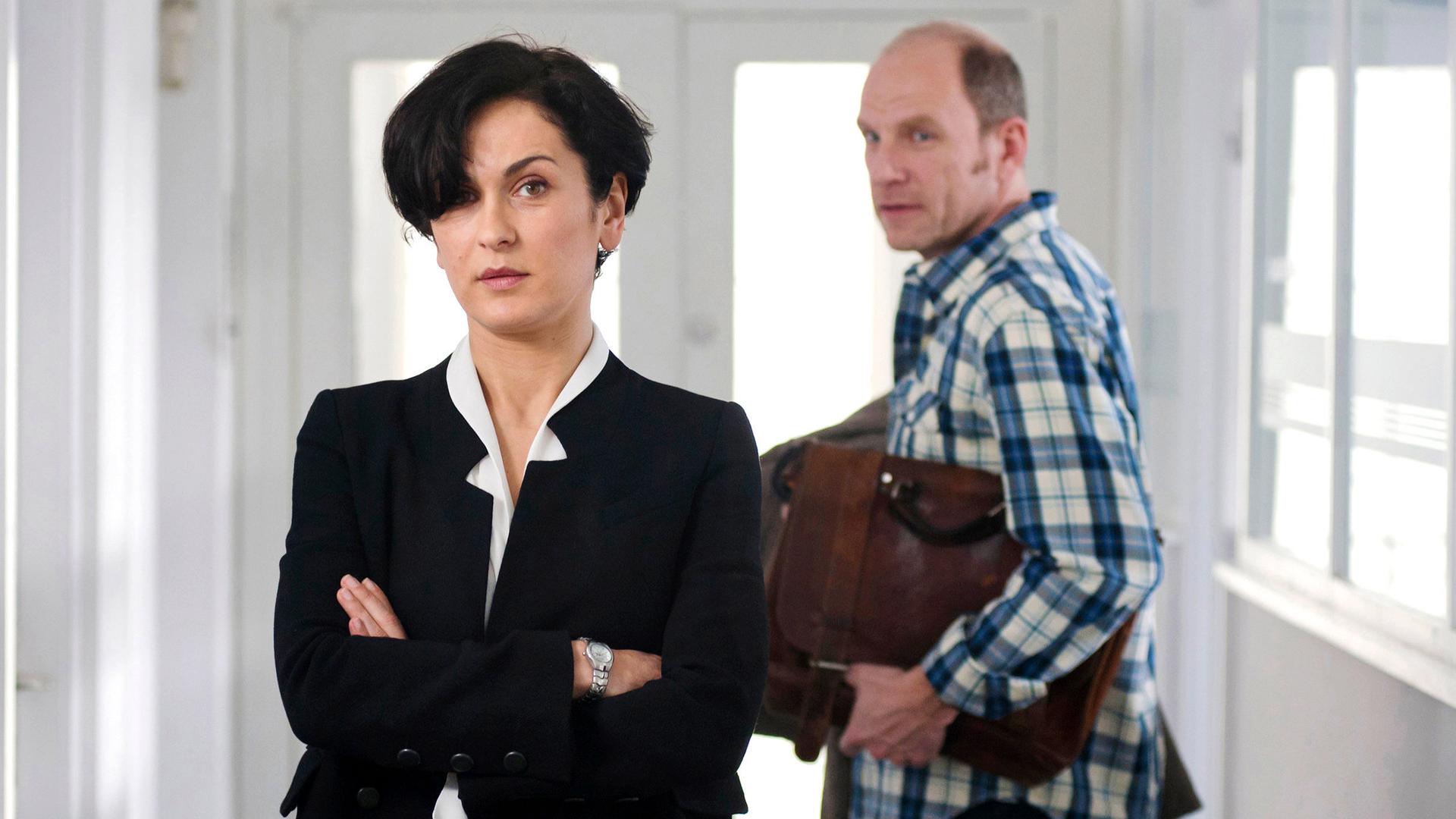 Hanna (Marie-Lou Sellem) steht mit verschränkten Armen im Anzug im Flur. Ihr Mann Martin (Götz Schubert) geht gerade zur Tür.