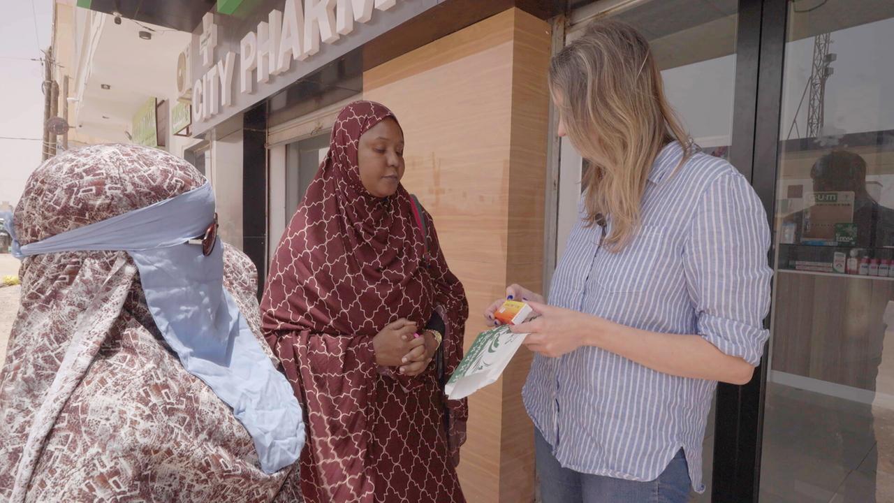 Das Bild zeigt eine Szene in einer urbanen Umgebung. Im Vordergrund stehen drei Frauen. Eine Frau auf der rechten Seite hat lange, blonden Haare und trägt ein kariertes Hemd. Sie hält ein weißes Paket in der Hand und scheint mit einer anderen Frau zu sprechen.  Die Frau in der Mitte trägt ein braunes, gemustertes Gewand und hat ihre Hände vor dem Körper verschränkt. Sie wirkt aufmerksam und interessiert an dem Gespräch. Die dritte Frau, auf der linken Seite, trägt ein hellblaues Tuch und ein graues Gewand, das ebenfalls traditionell ist.   Im Hintergrund ist ein Schaufenster eines Geschäfts zu sehen, auf dem "CITY PHARMACY" steht. Der Boden ist hell und die Umgebung scheint sonnig zu sein. Die Szene vermittelt eine Atmosphäre des Austauschs, möglicherweise über wichtige Themen der Frauen in ihrer Kultur.