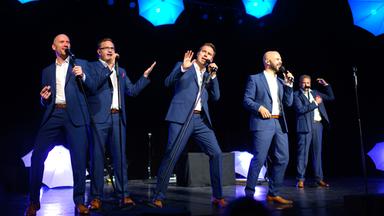 Mehrere Männer in blaugrauen Anzügen singen auf einer blau beleuchteten Bühne in Mikrofone.