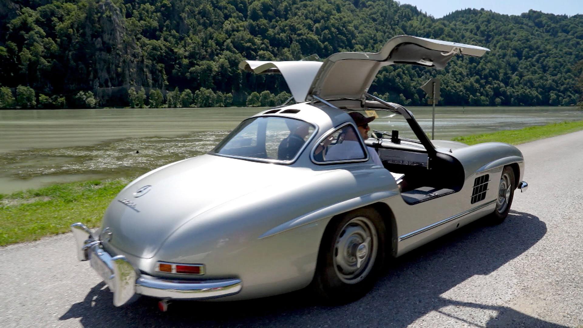 Das Bild zeigt einen klassischen Mercedes-Benz 300SL „Flügeltürer“, der auf einer kurvenreichen Landstraße fährt. Das Auto hat eine auffällige silberne Farbgebung und charakteristische Flügeltüren, die nach oben aufklappen. Im Hintergrund verläuft ein stiller Fluss, umgeben von einem bewaldeten Ufer mit grünen Hügeln.   Die Sonne scheint auf die Szenerie und es ist ein klarer, blauer Himmel zu sehen. Auf der Straße neben dem Auto befindet sich eine kleine Leitplanke sowie ein Straßenschild. Das Bild vermittelt eine Atmosphäre von Freiheit und Nostalgie, passend zu den Ausfahrten von Oldtimer-Liebhabern, die in der Beschreibung erwähnt werden.