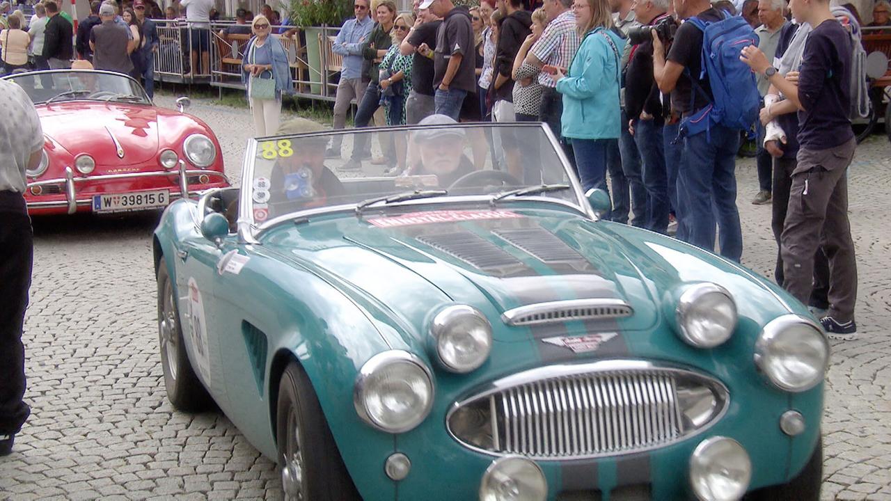 Das Bild zeigt eine belebte Straßenszene während eines Oldtimertreffens. Im Vordergrund steht ein teal-farbenes Austin Healey 3000 MK II Cabrio aus dem Jahr 1961. Das Auto hat runde Scheinwerfer und eine markante Kühlergrillgestaltung. Auf der Straße ist ein rotes klassisches Fahrzeug, möglicherweise ein Sportwagen, teilweise sichtbar.  Im Hintergrund sind viele Menschen zu sehen, die das Event beobachten. Einige Personen stehen auf einem Gehweg, während andere sich zu einem Geländer hinunterlehnen. Die Umgebung hat eine entspannte, festliche Atmosphäre. Die Veranstaltungsdetails deuten darauf hin, dass dies während einer Oldtimer-Rallye oder -Ausfahrt stattfindet. Die auf dem Asphalt verlegten Steine sind gut erkennbar und verleihen der Szenerie zusätzlichen Charme.