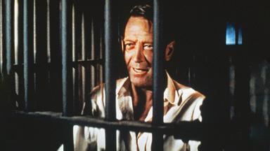 Alvarez Kelly ist mit weißem Hemd im Close-Up hinter schwarzen Gefängnisgittern zu sehen. Trotz seiner Situation und Zeichen der Müdigkeit im Gesicht lächelt er.