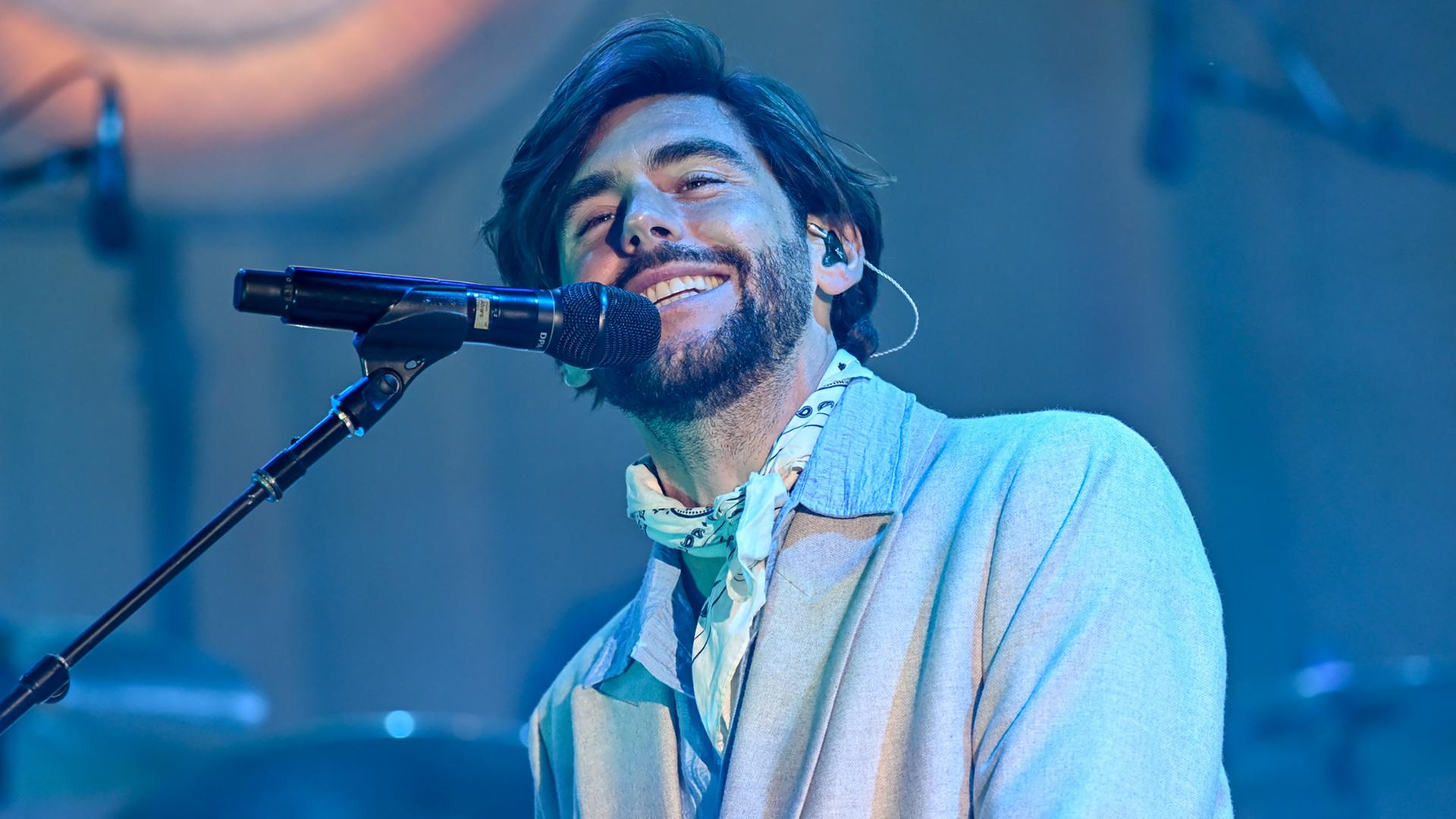 Der singende Alvaro Soler sitzt im Jackett und mit Halstuch vor einem Mikrofon im bläulichen Licht auf der Bühne.