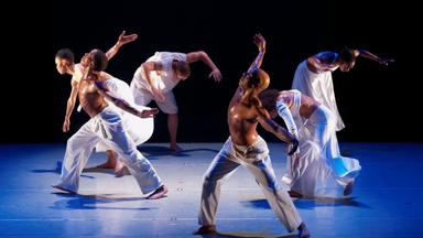Ensemble von 6 dunkelhäutigen Tänzern (3 female 3 masc.) in weißer Kleidung in ausdrucksstarken Bewegungsposen. Z.T. Oberkörper abgeknickt. Blick zu Boden oder Blick himmelwärts