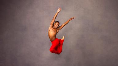 Dunkelhäutiger Tänzer in roter Hose, oberkörperfrei in extrem hohem Luftsprung. Im Sprung ein Bein zum Po angewinkelt. Zweites Bein an Knie des ersten angewinkelt. Arme nach oben gestreckt