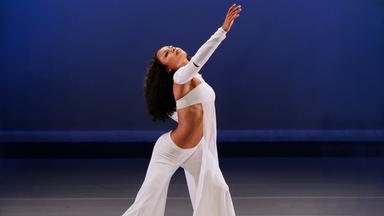 Dunkelhäutige Frau in weißer, weiter Hose, Bustier und Tuch. Bauchfrei. Seitenansicht. Ein Arm und Gesicht himmelwärts. Durchgebogener Körper -Hohlkreuz. Beine leicht auseinander