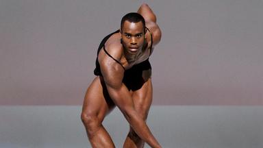 dunkelhäutiger muskulöser Solotänzer. Frontansicht. Durchdringender Blick. 3. Position des klassischen Balletts als Fußposition. Oberkörper nach vorne abgeknickt. Ein Arm nach vorne Richtung Boden