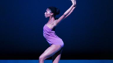 Tänzerin dunkelhäutig in Profil- bzw. Seitenansicht. Fliederfarbener Body. Blauer Bühnenboden, dunkelblauer Hintergrund. Körper im Hohlkreuz durchgebogen. Arme parallel gerade nach hinten gestreckt. Hinteres Bein leicht eingeknickt. Vorderes Bein auf angedeuteter Spitze