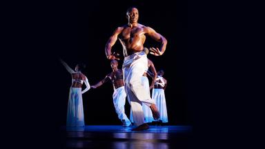 5 dunkelhäutige Tänzer vor schwarzem Hintergrund in weißer Kleidung. Im Vordergrund Tänzer in angedeuteter eingeleiteter Laufschritt-Sprungposition. Oberkörperfrei und lange weiße Hose. 4 Tänzer im Hintergrund z.T. in Rückenansicht